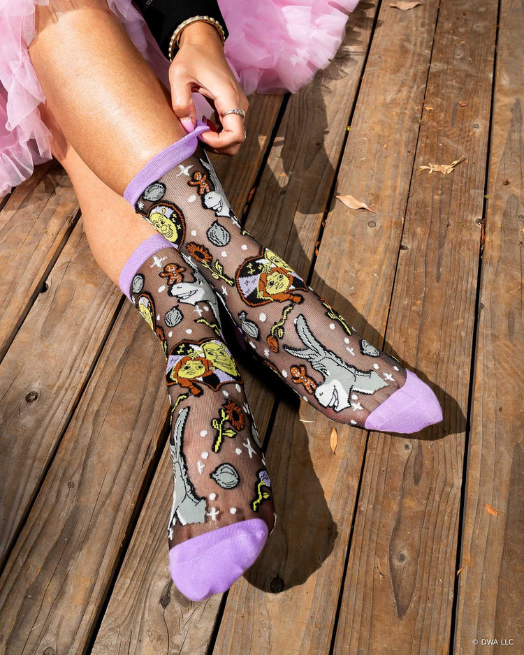 A woman's pair of legs wearing a pair of Shrek black sheer fashion socks designed by Sock Candy, sitting on a wooden stage.