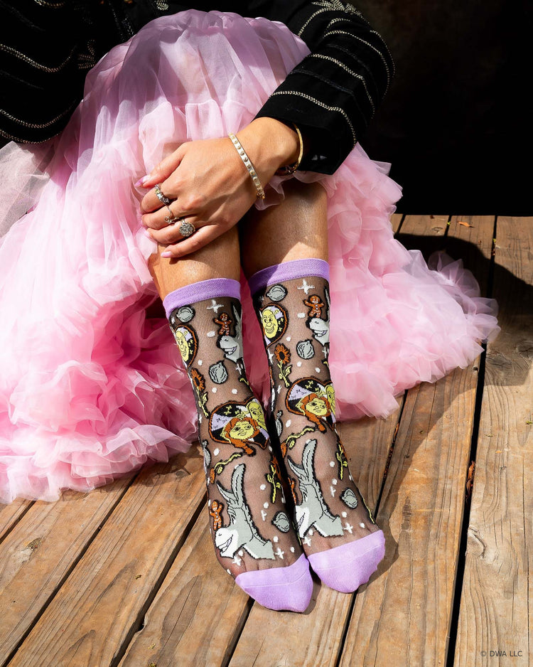 A woman seated on a wooden stage wearing Shrek black sheer fashion socks featuring Donkey, Gingy, and Shrek and Fiona, paired with a pink petticoat skirt.