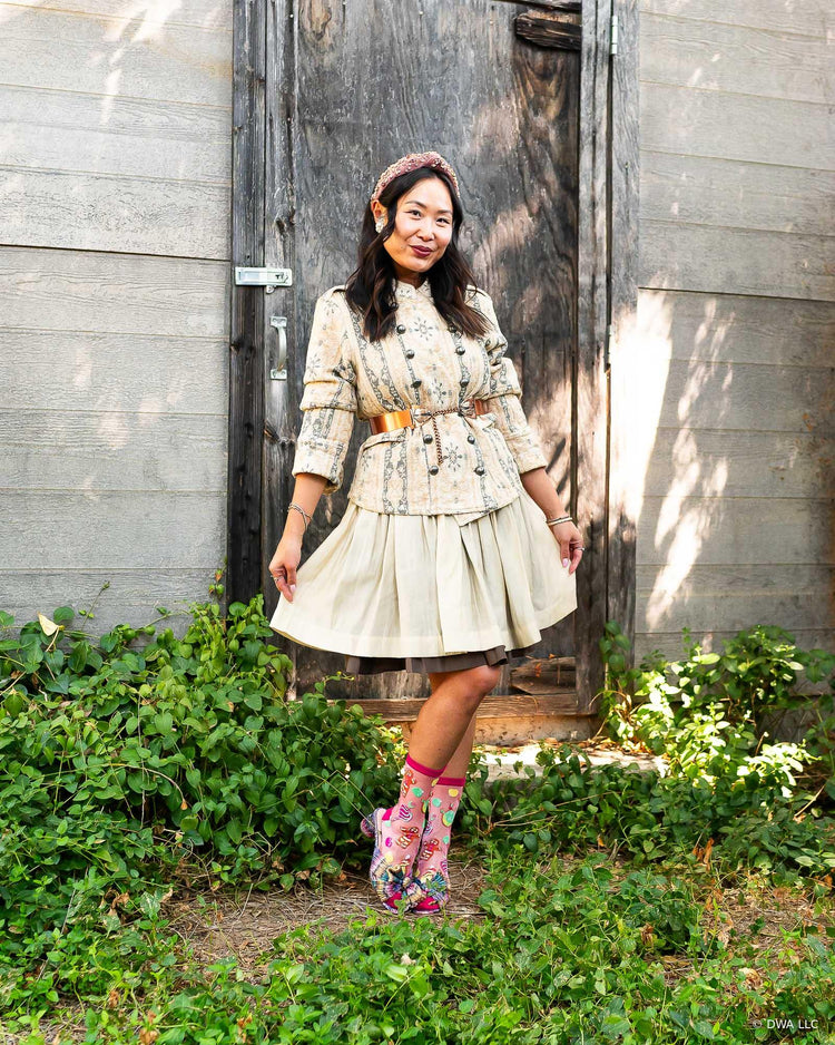 A woman stands outdoors in front of a weathered wooden door and gray siding, wearing a cream floral patterned belted jacket and pleated skirt. The vintage feminine style outfit is paired with sheer pink Shrek socks and metallic bow heels.