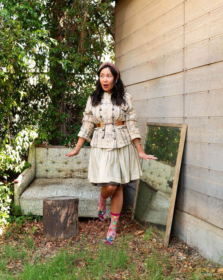 A woman stands outdoors in front of a an old tufted sofa, styled in a vintage feminine style with a floral jacket, Shrek sheer pink Gingy socks and metallic bow heels.