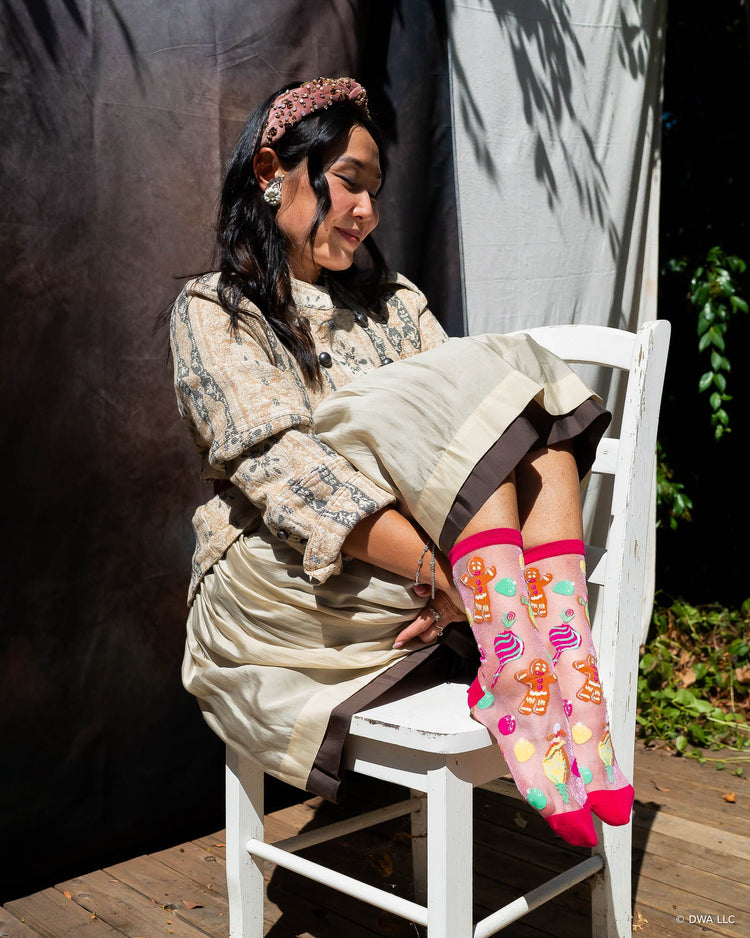 A woman sitting on a white wooden chair styled in a vintage feminine style with a floral jacket and wearing Shrek pink shimmer sheer Gingy socks with colorful candy designs.