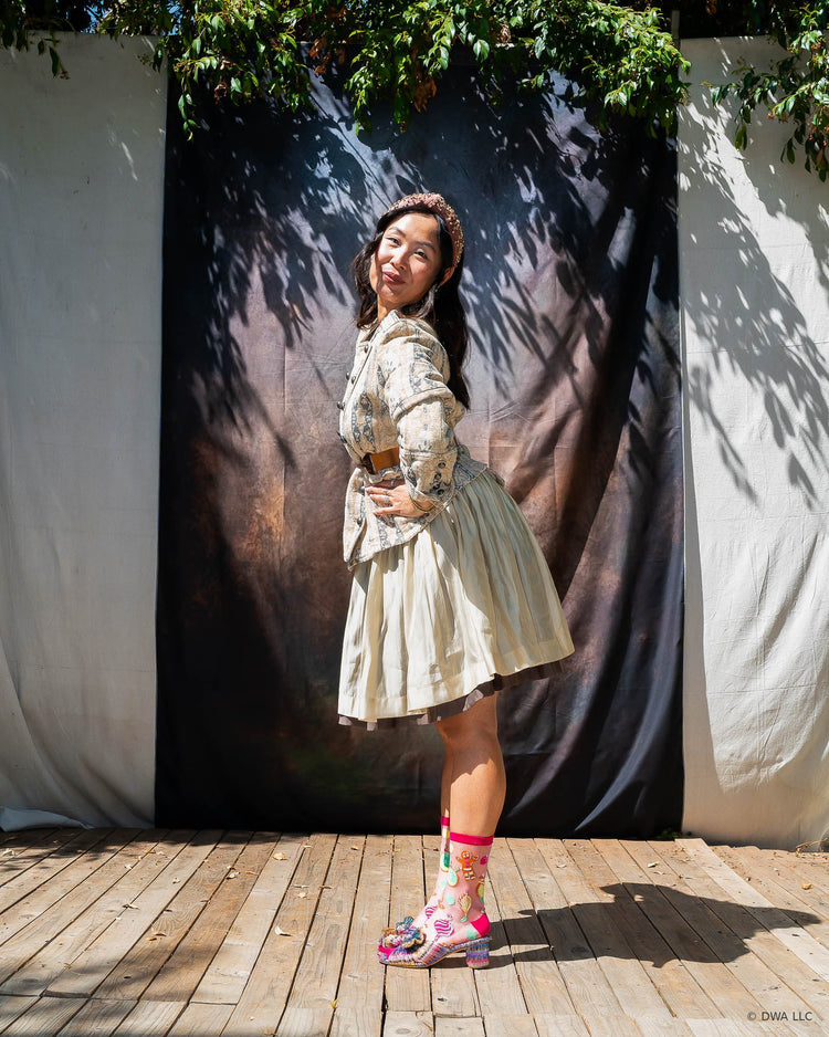 A woman posing outdoors on a wooden stage in front of a dark backdrop with tree shadows. She is styled in a vintage feminine way with a floral jacket, pleated skirt, pink shimmer sheer socks, and metallic bow heels.