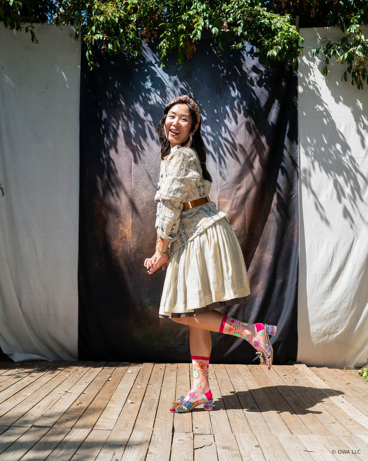 A woman posing on a wooden stage in front of a dark backdrop wearing sheer pink Shrek socks with metallic bow heels. She is styled in a vintage feminine way, with a floral patterned jacket and cream pleated skirt.