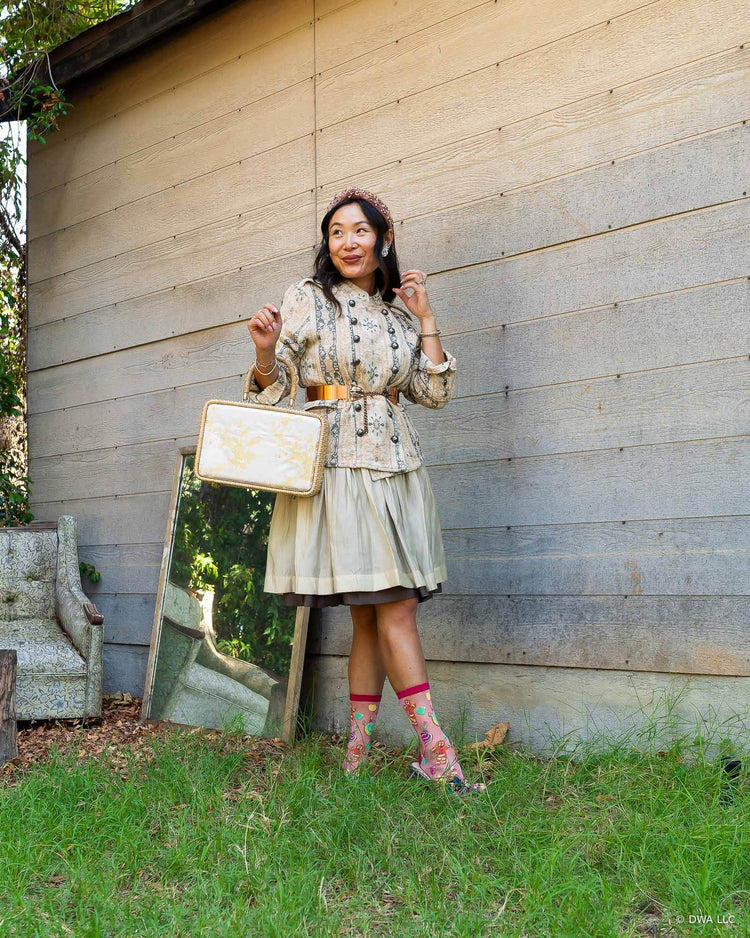 A woman stands outdoors against a weathered wooden building, wearing a vintage feminine style floral jacket belted at the waist, holding a matching cream-colored handbag. She is also wearing pink shimmer sheer patterned socks and metallic bow heels, with a small mirror leaning against the wall beside her.
