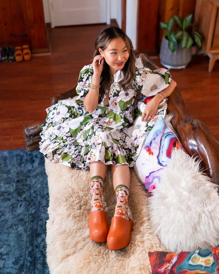 A woman sitting on a couch wearing a floral-patterned dress and sheer Sock Candy socks in a fox motif paired with tan clogs.